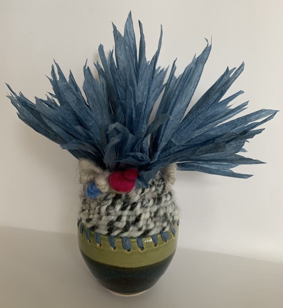 Ceramic vase base with grey weaving above and topped with strips of blue paper protruding from the top of the yarn